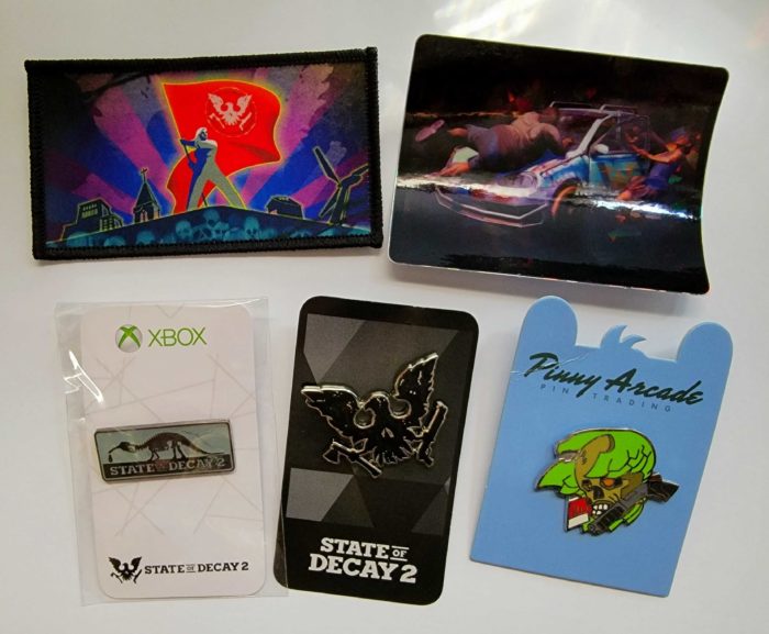 Image featuring an achievement patch (with survivor waving a red flag), a holographic sticker of a zombie attacking a car, a Bronto Gas and State of Decay logo pin, a skeagle logo pin and a red talon pin.